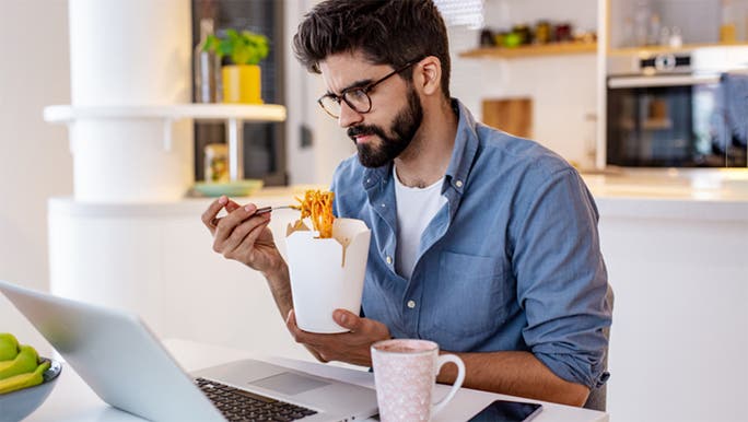 A man with dark hair and a beard is eating from a takeaway container while he works on a laptop